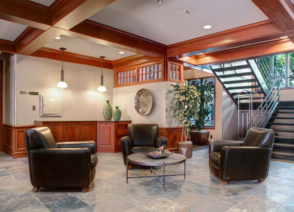 Cozy lobby here at Cheyenne Creek Apartment Homes with four black leather chairs around a round coffee table. The room has tile flooring, wood accents, a staircase, and a large potted plant.