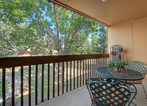 Cozy balcony here at Eagle Ridge Apartment Homes with metal table, chairs, and potted plants overlooking lush trees. Sunlight filters through branches, creating a serene atmosphere.