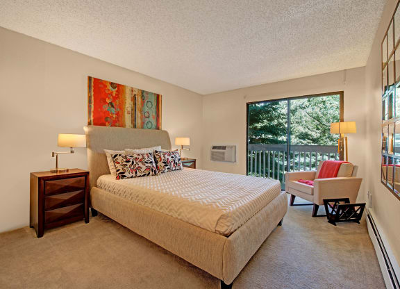 Contemporary bedroom here at Eagle Ridge Apartment Homes with a beige bed, colorful abstract art, and large window overlooking trees. Warm lighting and a cozy armchair add comfort.