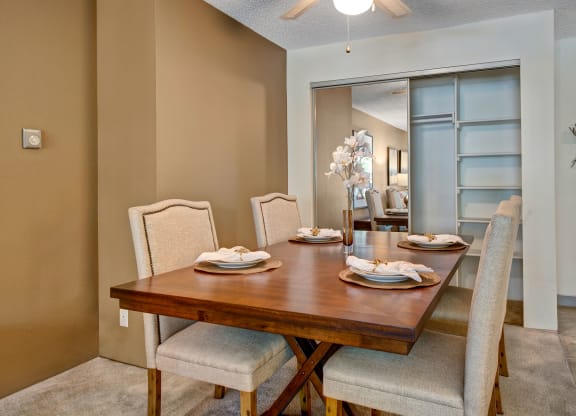 Cozy dining room here at Eagle Ridge Apartment Homes with a wooden table set for four. Beige upholstered chairs surround the table, which is decorated with plates, napkins, and a vase of white flowers. Neutral tones create a warm, inviting atmosphere.