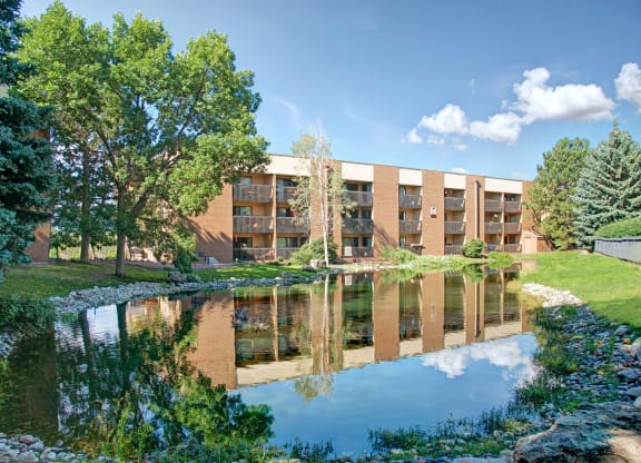 A serene pond here at Eagle Ridge Apartment Homes reflects a four-story brick apartment building surrounded by lush green trees under a blue sky with fluffy clouds, conveying tranquility.