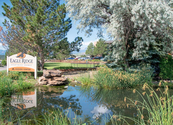 A sign for "Eagle Ridge Apartment Homes" stands amid lush greenery next to a tranquil pond with a stone fountain. The scene conveys serenity.