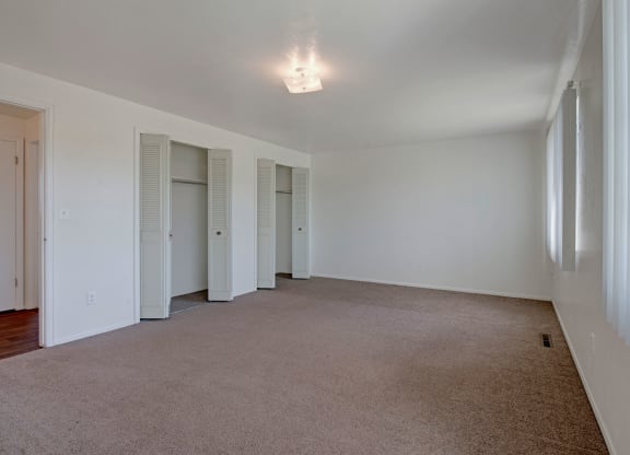 Spacious empty room here at Yorkshire Square Apartment Homes with beige carpet, neutral walls, and a ceiling light. Features include two open closets with louvered doors and large windows with vertical blinds, creating a serene atmosphere.
