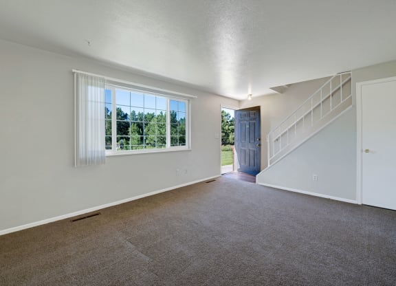 Empty living room here at Yorkshire Square Apartment Homes with beige carpet, white walls, and a large window showing trees outside. An open blue door and staircase add depth and light.