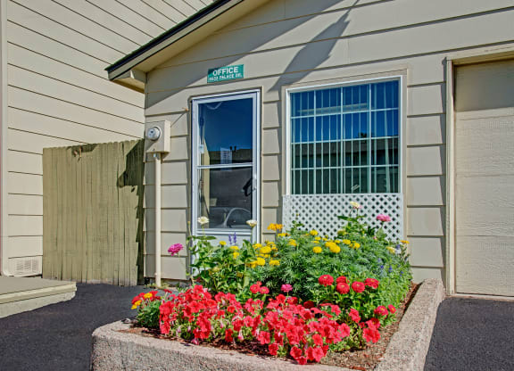 A small beige leasing office here at Yorkshire Square Apartment Homes with flowers in front, including red and yellow blooms. Bright, sunny day with a clear blue sky. The mood is cheerful.