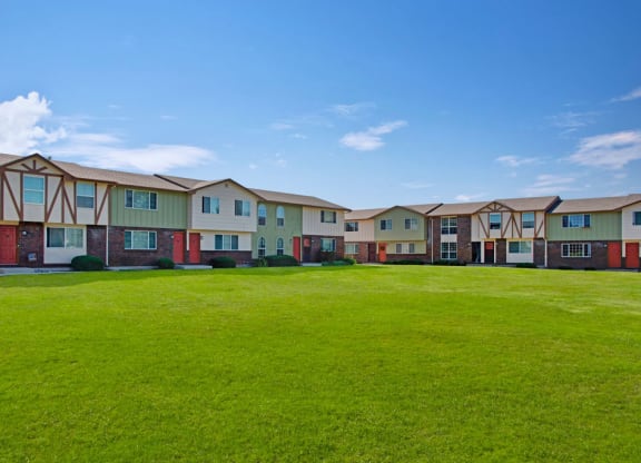 Row of suburban Yorkshire Square Apartment Homes with red doors and Tudor-style accents, surrounded by a large, well-kept grassy lawn under a clear blue sky.