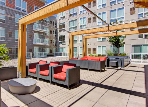 Modern apartment courtyard here at 7west Apartment Homes with gray buildings, featuring wooden pergolas and red-cushioned patio furniture. Bright, sunny atmosphere with a relaxed vibe.