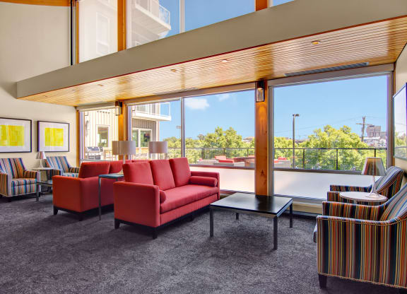 Spacious modern lounge here at 7west Apartment Homes with large windows, bright red couches, striped chairs, and abstract yellow art. Natural light creates a vibrant, welcoming atmosphere.