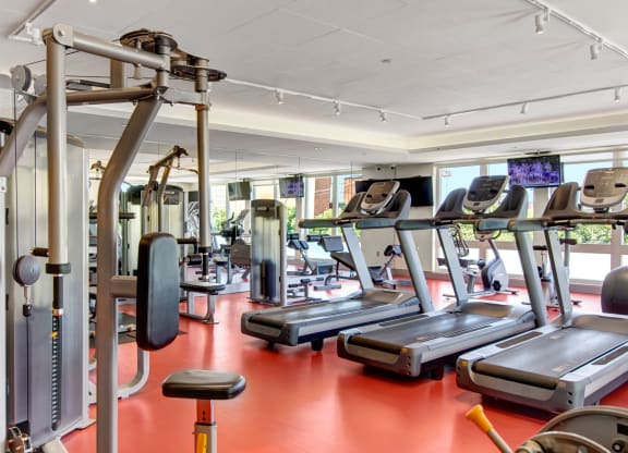 Modern gym here at 7west Apartment Homes with red flooring, featuring treadmills, weight machines, and large windows allowing natural light. Energetic and inviting atmosphere.