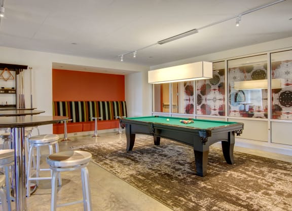 Modern recreation room here at 7west Apartment Homes with a pool table on a patterned rug. There's a seating area with striped cushions, high tables with stools, and abstract wall art.