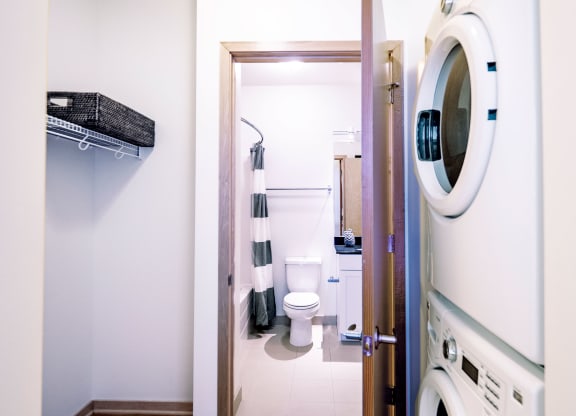 Laundry room here at 7west Apartment Homes with a stacked washer and dryer on the right, and a door opening into a bathroom with a toilet, sink, and striped shower curtain in neutral tones.