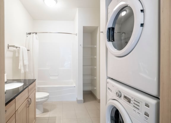 Bright bathroom here at 7west Apartment Homes with a bathtub, toilet, and wooden vanity. A stacked washer and dryer are on the right. Shelving is visible in the wall niche. Minimalist and clean.