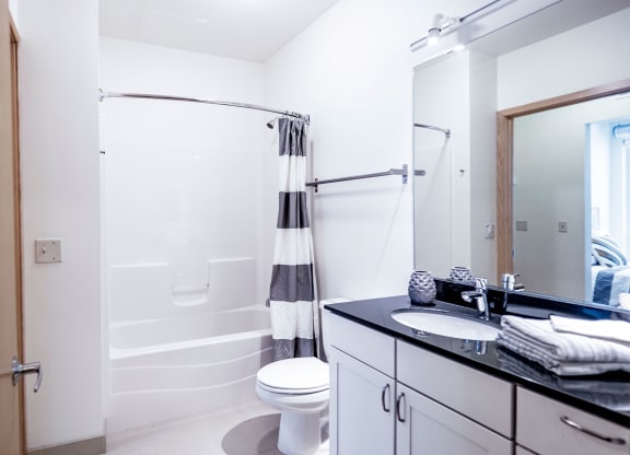 Bright, modern bathroom here at 7west Apartment Homes with white walls, a bathtub with a striped curtain, and a black countertop sink. Clean, minimalist design conveys tranquility.