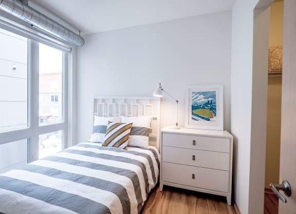 Bright, minimalistic bedroom here at 7west Apartment Homes with a window, single bed with striped bedding, and a colorful pillow. A white dresser holds a lamp and art print.