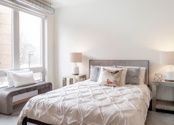 Bright bedroom here at 7west Apartment Homes with a neatly made bed, featuring white and gray linens and a decorative bird pillow. Soft lighting from two lamps creates a cozy ambiance.