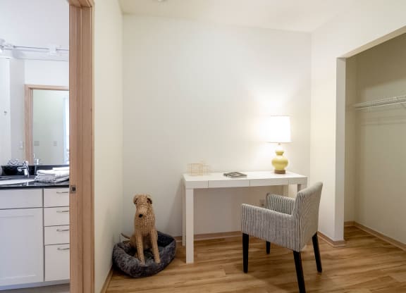 A minimalist room here at 7west Apartment Homes with wooden flooring features a white desk with a lamp, a striped chair, and a woven dog in a bed. An open closet and bathroom are visible.