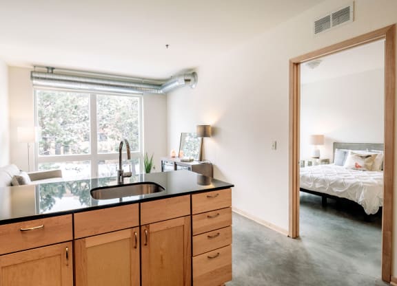 Modern apartment interior here at 7west Apartment Homes with an open kitchen, featuring wooden cabinets and a black countertop. Bright living area with a window and a glimpse of a cozy bedroom.