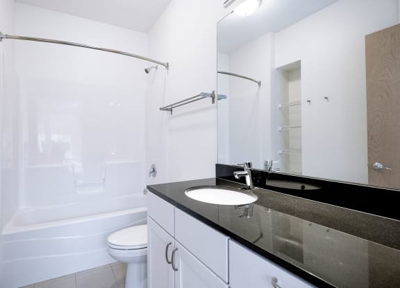Modern bathroom here at 7west Apartment Homes with white walls and fittings. Features a bathtub with a shower curtain, a toilet, and a sleek black countertop with a sink.