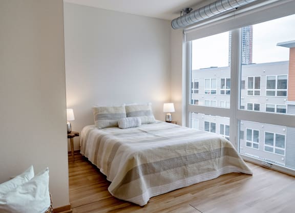 A modern bedroom here at 7west Apartment Homes with a neatly made bed featuring striped bedding. Two lamps on bedside tables provide soft lighting. Large windows reveal a city view.