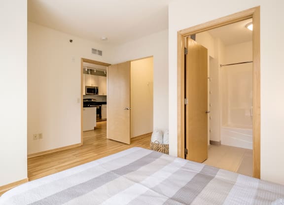 A bright bedroom here at 7west Apartment Homes featuring a neatly made bed, light wood floor, and cream walls. Open doors reveal a modern kitchen and a bathroom, creating a cozy, inviting ambiance.