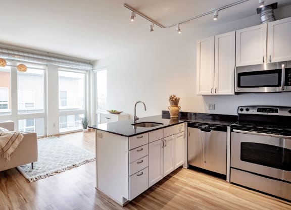 Modern apartment interior here at 7west Apartment Homes with a bright open-plan living room and kitchen, featuring large windows, light wood floors, a beige sofa, and sleek appliances.