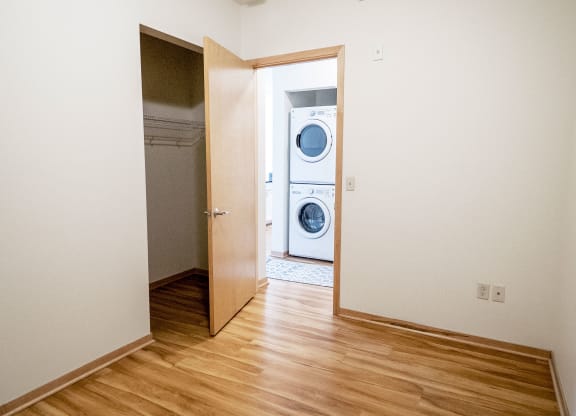 Empty room here at 7west Apartment Homes with light wood flooring, open door leading to a closet, and view of stacked washer and dryer in adjoining room. Bright and minimalistic.
