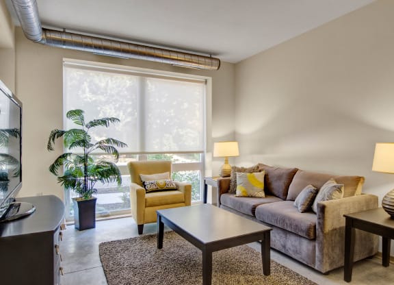 A cozy living room here at 7west Apartment Homes with a brown sofa and yellow armchair next to large windows. Warm lighting from lamps and greenery create a relaxed, inviting atmosphere.