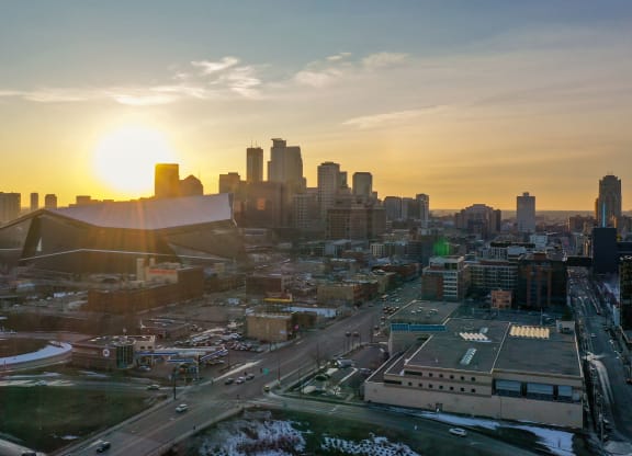 Sunset over a city skyline at 7west Apartment Homes with a prominent, modern stadium. The sun casts a warm glow, highlighting tall buildings and snow-dusted streets below.
