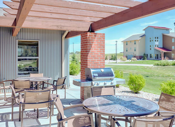 Outdoor patio here at Dakota Ridge Apartment Homes with a wooden pergola, metal tables, chairs, and a grill beside a brick column. Nearby, a lawn leads to a modern multi-story building. Bright and inviting atmosphere.