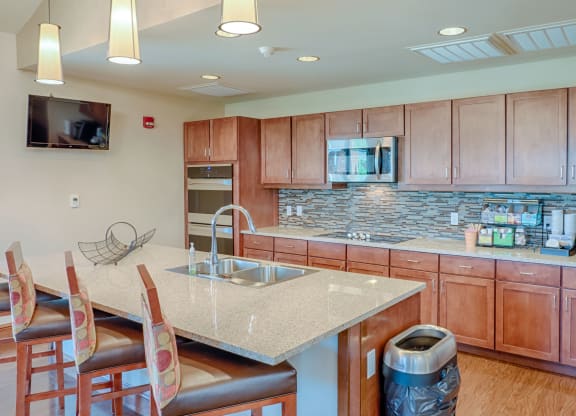 Modern kitchen here at Dakota Ridge Apartment Homes with wooden cabinets, tiled backsplash, and stainless steel appliances. Granite island counter has three chairs and a fruit basket.