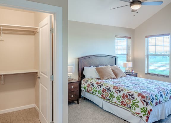 Cozy bedroom here at Dakota Ridge Apartment Homes with a colorful floral bedspread, wooden headboard, and bedside table. Open closet with shelves. Warm light streams through large windows.