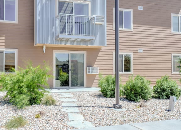 Two-story building exterior here at Dakota Ridge Apartment Homes with brown siding and metal balcony. A path of flat stones leads to a glass door, surrounded by green shrubs and pebbles.