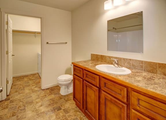 Spacious bathroom here at Phoenix Ridge Apartment Homes with wood vanity, marble countertop, and mirror. Toilet beside a door leading to laundry room. Warm, clean, and functional atmosphere.
