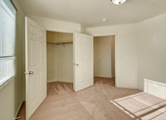 Empty beige bedroom here at Phoenix Ridge Apartment Homes with soft carpet, an open closet with double doors, and sunlight streaming through window blinds, casting shadows on the floor.