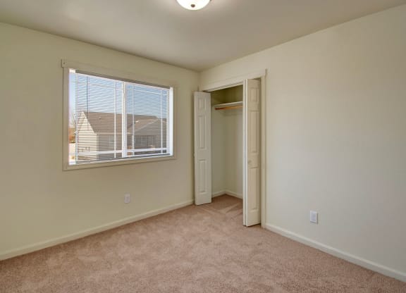 Empty bedroom here at Phoenix Ridge Apartment Homes with beige carpet, cream walls, and a window with blinds. An open closet with a white door is on the right, under a ceiling light.