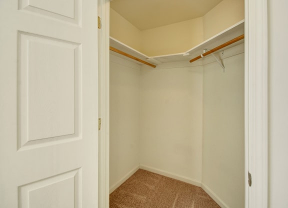 Empty walk-in closet here at Phoenix Ridge Apartment Homes with beige walls, carpeted floor, and wooden hanging rods on white shelves. The open door reveals a spacious, clean feel.