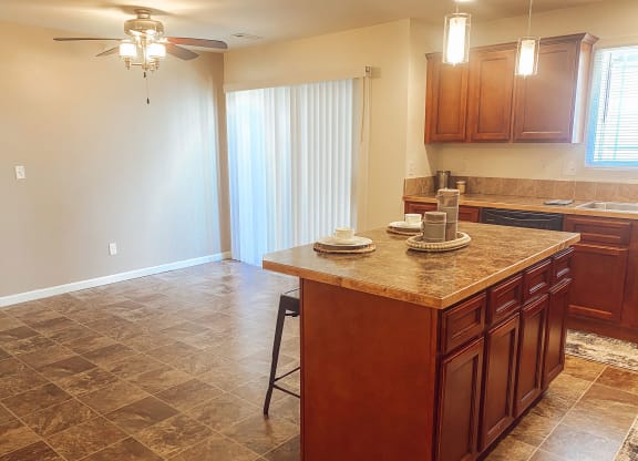 Warm kitchen here at Phoenix Ridge Apartment Homes with brown cabinets and tile floor. An island with dishes and stools. Soft lighting from ceiling fixtures creates a cozy atmosphere.