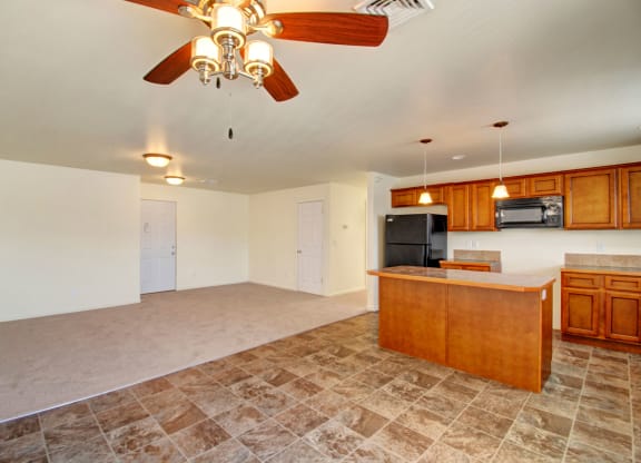 Open-concept room here at Phoenix Ridge Apartment Homes with beige walls and dual flooring: carpeted area leads to a tiled kitchen with wooden cabinets, an island, and a ceiling fan. Cozy and spacious.