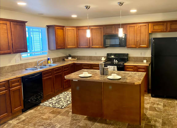 Spacious kitchen here at Phoenix Ridge Apartment Homes with warm wooden cabinets, black appliances, and a large island. Bright lighting creates a welcoming, cozy atmosphere.