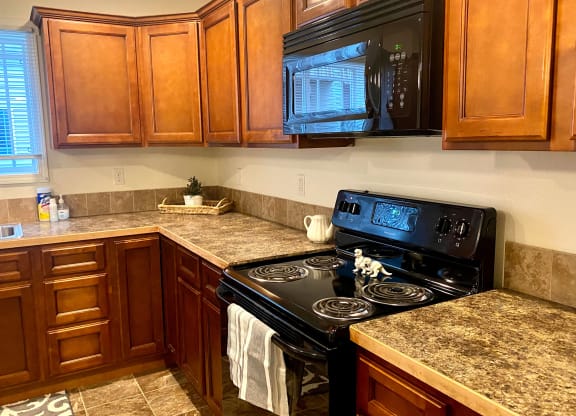 A cozy kitchen here at Phoenix Ridge Apartment Homes with warm wooden cabinets, granite countertops, a black stove with a striped towel, and a plant by a bright window on a tiled floor.