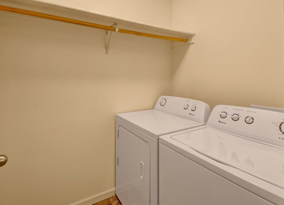 Small laundry room here at Phoenix Ridge Apartment Homes with white washer and dryer against a beige wall. A wooden shelf with a hanging rod is above the appliances. The space is tidy and functional.
