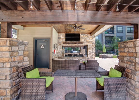 Covered patio area here at the Enclave at Brookside Apartments with wicker furniture and lime green cushions. Stone walls and wooden beams complement a TV above a fireplace. Relaxed, inviting atmosphere.