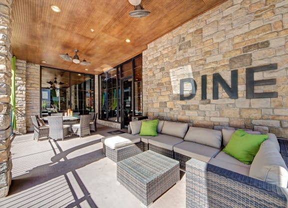 Outdoor patio here at the Enclave at Brookside Apartments with wicker furniture and green cushions, under a wooden ceiling. Stone wall with "DINE" sign. Sunny, inviting atmosphere.
