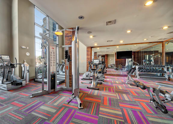 Bright, spacious gym here at the Enclave at Brookside Apartments with vibrant multicolored carpet, modern exercise equipment including ellipticals and weights. Large windows provide natural light.