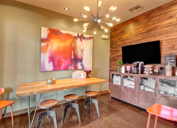 Modern coffee room here at the Enclave at Brookside Apartments with a wooden table, three stools, and orange chairs. Abstract red-and-white art adorns the wall. A cabinet holds coffee supplies. Warm lighting and a cozy atmosphere.