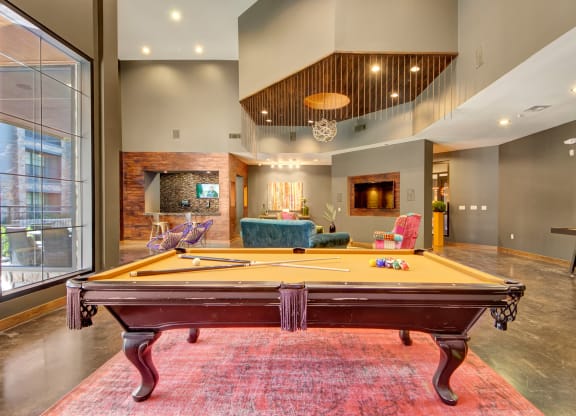 Spacious lounge here at the Enclave at Brookside Apartments with a central pool table on a red rug. Modern decor, high ceilings, large windows, comfortable seating, and a cozy atmosphere.