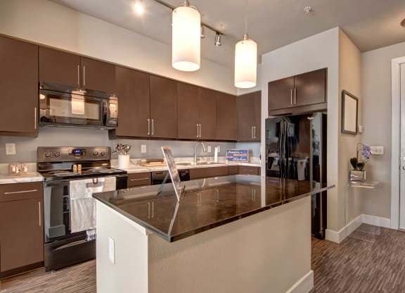 Modern kitchen here at the Enclave at Brookside Apartments with dark wood cabinets, black appliances, and granite countertops. Two pendant lights hang above a central island. Light and minimalist tone.