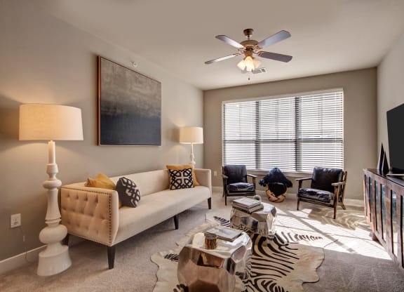 Modern living room here at the Enclave at Brookside Apartments with neutral tones featuring a beige sofa, patterned cushions, abstract wall art, zebra rug, metal tables, and a window with blinds.
