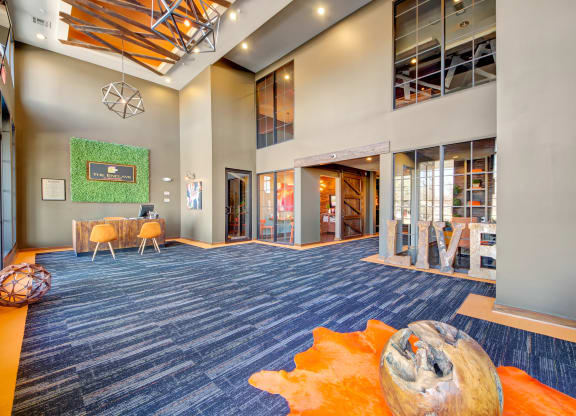 Spacious lobby with high ceilings here at the Enclave at Brookside Apartments, modern geometric lighting, and a mix of blue carpet and orange accents. Wooden decor pieces and casual seating create a welcoming atmosphere.