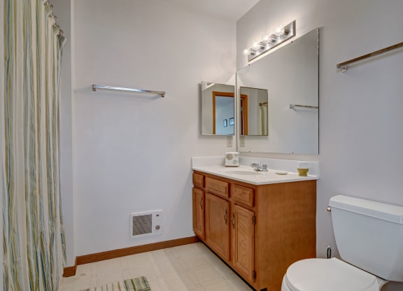 Small bathroom with a white toilet, wooden vanity, and large mirror. Light fixture above, and striped shower curtain to the left. Calm, minimal look. Arbor Ridge Apartments